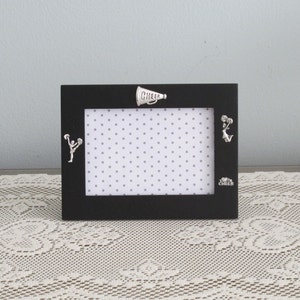 May include: Black picture frame with a white polka dot background. The frame has three silver charms attached to it: a cheerleader, a megaphone with the word "CHEER" on it, and a heart with the word "CHEER" on it.
