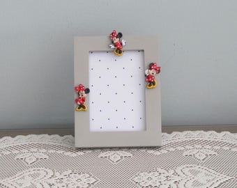 May include: A rectangular light gray picture frame featuring three small, colorful Minnie Mouse character embellishments attached to the top and side borders. The frame surrounds a white insert decorated with a pattern of small black dots.