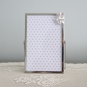 May include: A silver picture frame with a white background and a silver decorative bow on the top right corner. The background has a small white polka dot pattern.