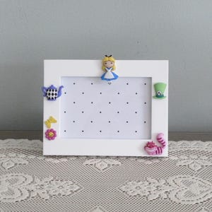 May include: A white rectangular earring display frame featuring a perforated white board with black dots. The frame is decorated with small character-themed embellishments including a girl, a teapot, a green hat, a yellow butterfly, a flower, and a cat.