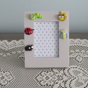 May include: A white picture frame with a polka dot background. The frame has five small, colorful, 3D bug decorations on the front: a green caterpillar, a red ladybug, a black ant, a yellow bee, and a blue and yellow dragonfly.