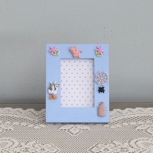 May include: A light blue picture frame with a white polka dot background. The frame is decorated with a pink pig, a white goose, a spider, a spider web, and two pink flowers.