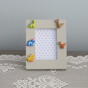 May include: A white picture frame with a polka dot background and five colorful dinosaur figurines attached to the frame.