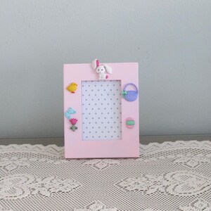 May include: A pink picture frame with a white bunny, a yellow bird, a purple purse, a butterfly, and a flower. The frame has a white background with gray polka dots.