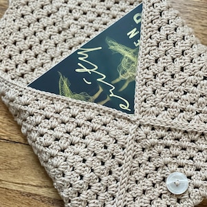May include: A beige crocheted book sleeve with a white button. The sleeve is folded over a book with a dark blue cover and the text "Once Upon a Broken Heart" in white.