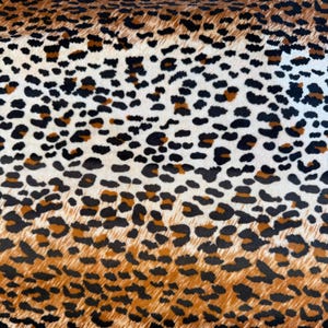 May include: Close-up of a fabric with a leopard print pattern. The pattern features black, brown, and tan spots on a white background. The spots vary in size and shape, creating a naturalistic animal print design.