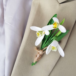 May include: A white and yellow flower boutonniere with green leaves and a brown twine bow. The boutonniere is pinned to a beige suit jacket.