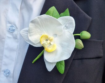 Puede incluir: Un boutonniere de orquídea blanca con hojas verdes, perfecto para una boda u ocasión especial.