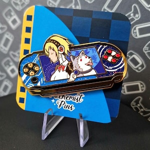 Puede incluir: Pin de esmalte de color dorado que representa una consola de videojuegos portátil azul y blanca con un personaje femenino y un perro blanco en la pantalla. El pin está en un expositor azul y blanco con el texto "Achemist Pins".