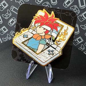 May include: A gold-colored enamel pin featuring a cartoon character with red hair and a blue shirt, emerging from a white handheld gaming device with a screen showing a landscape. The device has a gold-colored border and flames on the side. The pin is on a black background.