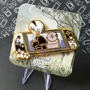 Puede incluir: Pin de esmalte de color dorado que representa a un personaje de videojuego sosteniendo una consola de juegos portátil. El personaje es una mujer con el pelo largo y blanco y un atuendo negro. La consola es una versión estilizada de la Nintendo Switch. El pin está colocado sobre un fondo de follaje verde y el texto "Alchemist Pins".