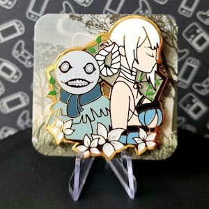 May include: A gold-tone enamel pin featuring a white-haired woman with a blue and white creature beside her. The pin is set against a green and brown background with white flowers.