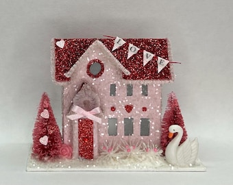 May include: A decorative miniature house in light pink with a red glitter roof, featuring a banner that spells LOVE, a red glitter door with a pink bow, two pink bottle brush trees with heart accents, and a white swan figurine on a base.