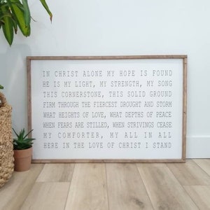 May include: A wooden sign with a white background and black text. The text reads "In Christ alone my hope is found, He is my light, my strength, my song, This cornerstone, this solid ground, Firm through the fiercest drought and storm, What heights of love, what depths of peace, When fears are stilled, when strivings cease, My comforter, my all in all, Here in the love of Christ I stand."