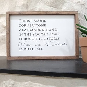 Pode incluir: Uma placa de madeira com fundo branco e texto preto. O texto diz "CHRIST ALONE CORNERSTONE WEAK MADE STRONG IN THE SAVIOR'S LOVE THROUGH THE STORM He is Lord LORD OF ALL".