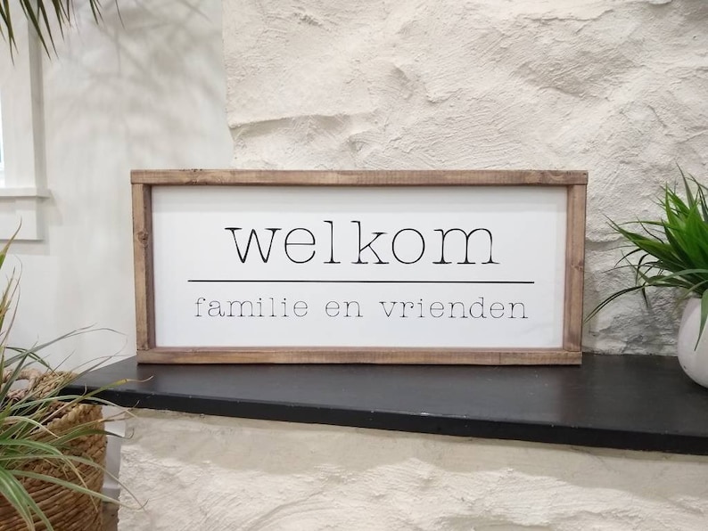 May include: A rectangular wooden sign with the word "welkom" above "familie en vrienden" in black font on a white background. The sign has a dark brown wooden frame and is displayed on a black surface.