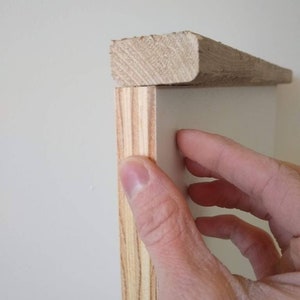 May include: Close-up of a wooden trim piece being installed. The trim is a light brown color and is being placed on top of a white surface. The wood grain is visible. The trim is rectangular with a rounded edge.