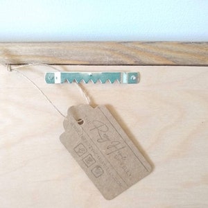 May include: A wooden panel with a metal sawtooth hanger attached. A brown paper tag is tied to the panel with twine. The tag has text and logos printed on it.
