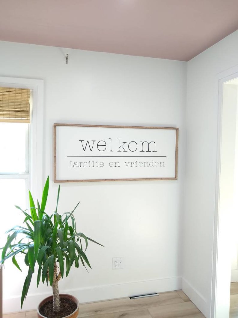 May include: A rectangular wooden framed sign with the word "Welkom" above the words "familie en vrienden" in a white interior. A potted plant is in the foreground. The ceiling is painted a light pink color.
