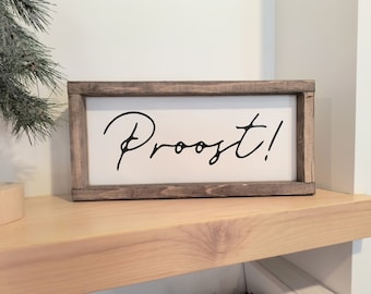 May include: A wooden sign with a white background and a black cursive inscription that reads "Proost!" The sign is framed in a dark brown wood frame.