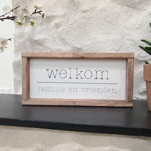 May include: A rectangular wooden sign with the word "welkom" above the words "familie en vrienden" in black font. The sign is set against a white background with a plant in a terracotta pot to the right.