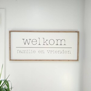 May include: A rectangular wooden sign with the word "welkom" above the words "familie en vrienden" in black font. The sign is framed in a light brown wood and hangs on a white wall.