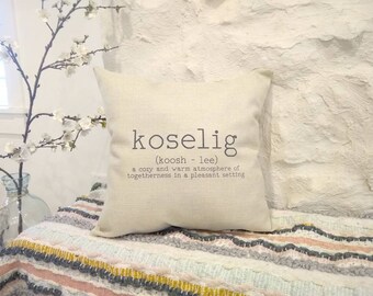 May include: A white throw pillow with the word "koselig" printed in black. The definition of the word is "a cozy and warm atmosphere of togetherness in a pleasant setting".