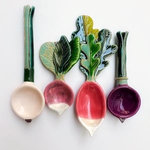 May include: Four ceramic spoons in the shape of vegetables. The spoons are in the shape of a radish, onion, and scallion. The radish spoons are red and white. The onion spoon is light pink. The scallion spoons are green and purple.