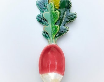 May include: A ceramic radish-shaped spoon with a vibrant design. The spoon features a bright red body with a white tip, and green, yellow, and blue leaves. The spoon is likely for kitchen or dining use.