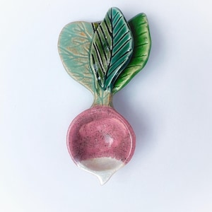 May include: A ceramic radish-shaped spoon rest. The spoon rest features a pink radish body with a white tip and green leaves. The leaves have black vein details and are in shades of green and teal.