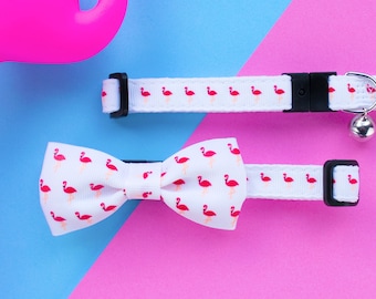 May include: A pink flamingo toy, a white bow tie with a flamingo print, and a white collar with a flamingo print and a silver bell.