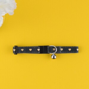 May include: A black cat collar with a gold heart pattern and a silver bell. The collar is on a yellow background.