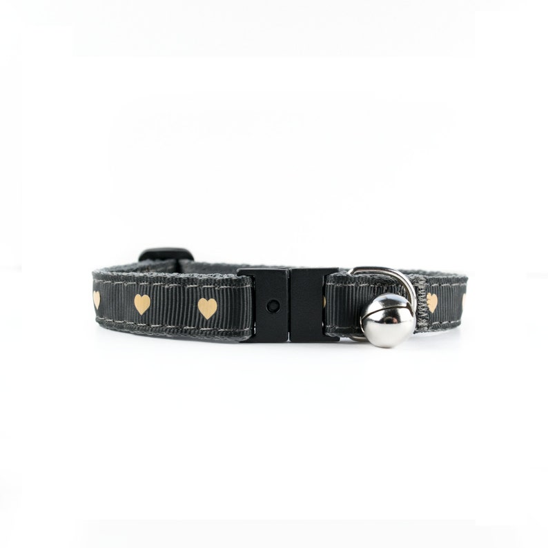 May include: A gray cat collar with a safety breakaway buckle and a small bell. The collar is decorated with gold hearts.