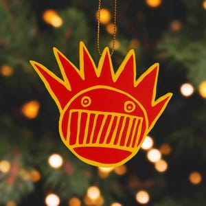 Puede incluir: Un adorno navideño rojo y amarillo con un diseño de cara de dibujos animados. El adorno cuelga frente a un fondo borroso de un árbol de Navidad con luces. El adorno tiene forma de sol con una cara.