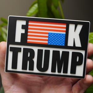 May include: Rectangular sticker with the text "F**K TRUMP" in bold black letters. The design incorporates the American flag, with red and white stripes and blue stars. The sticker has a white border.