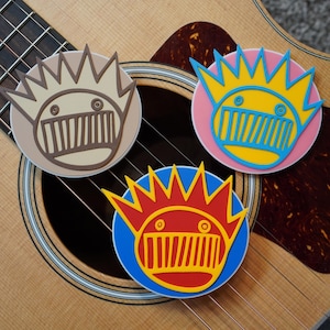 Könnte beinhalten: Drei bunte runde Aufkleber mit einem Cartoon-Kronen-Design, die auf einer Akustikgitarre liegen. Die Aufkleber zeigen ein Kronen- und Gesichtsdesign in verschiedenen Farbkombinationen: Beige und Braun, Gelb und Blau auf Rosa sowie Rot und Gelb auf Blau.