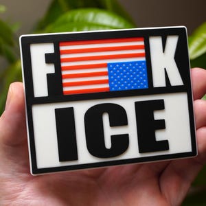 Może przedstawiać: Prostokątna naklejka z napisem "F**K ICE" pogrubionymi czarnymi literami. Litery "F" i "K" są oddzielone fragmentem amerykańskiej flagi. Naklejka jest trzymana w dłoni.