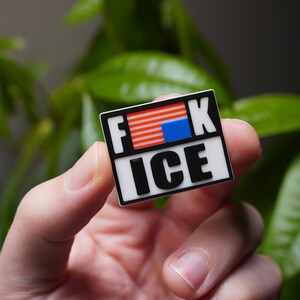 Może przedstawiać: Prostokątna przypinka z napisem "F**K ICE" pogrubionymi czarnymi literami na białym tle. G&oacute;rna część przedstawia stylizowany wz&oacute;r amerykańskiej flagi. Przypinka trzymana jest w dłoni na rozmytym zielonym tle.