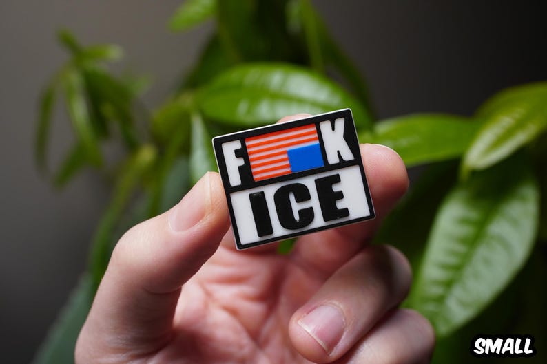 Może przedstawiać: Prostokątna przypinka z napisem "F**K ICE" pogrubionymi czarnymi literami na białym tle. G&oacute;rna część przedstawia stylizowany wz&oacute;r amerykańskiej flagi. Przypinka trzymana jest w dłoni na rozmytym zielonym tle.
