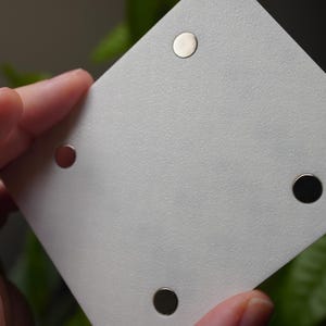 Może przedstawiać: Biała, kwadratowa karta z czterema małymi, okrągłymi magnesami. Magnesy są srebrne i czarne. Karta jest trzymana w dłoni, z rozmytym zielonym tłem.