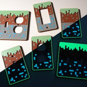 Könnte beinhalten: Eine Sammlung dekorativer Lichtschalter- und Steckdosenabdeckungen. Die Abdeckungen weisen ein pixeliges Design mit grünen, braunen, grauen und blauen Farben auf, das an eine Videospiel-Landschaft erinnert. Einige Abdeckungen leuchten im Dunkeln.