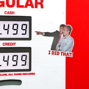 Op de afbeelding: Een benzinepompdisplay met prijzen van $4,499 per gallon voor contant en credit. Een sticker met twee mannen en de tekst "I DID THAT!" is op de pomp geplaatst. Het woord "REGULAR" is bovenaan zichtbaar.