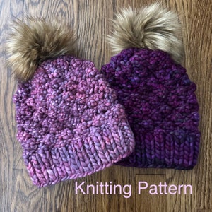 May include: Two hand-knitted beanies in shades of purple and pink, each topped with a fluffy, brown faux fur pom-pom. The beanies are made with thick, textured yarn. The text "Knitting Pattern" is visible at the bottom.