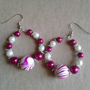 May include: A pair of hoop earrings made with pink and white beads. The earrings have a large, white, swirl-patterned bead on each side.
