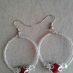 May include: A pair of silver hoop earrings with a red bead and clear crystal accents.