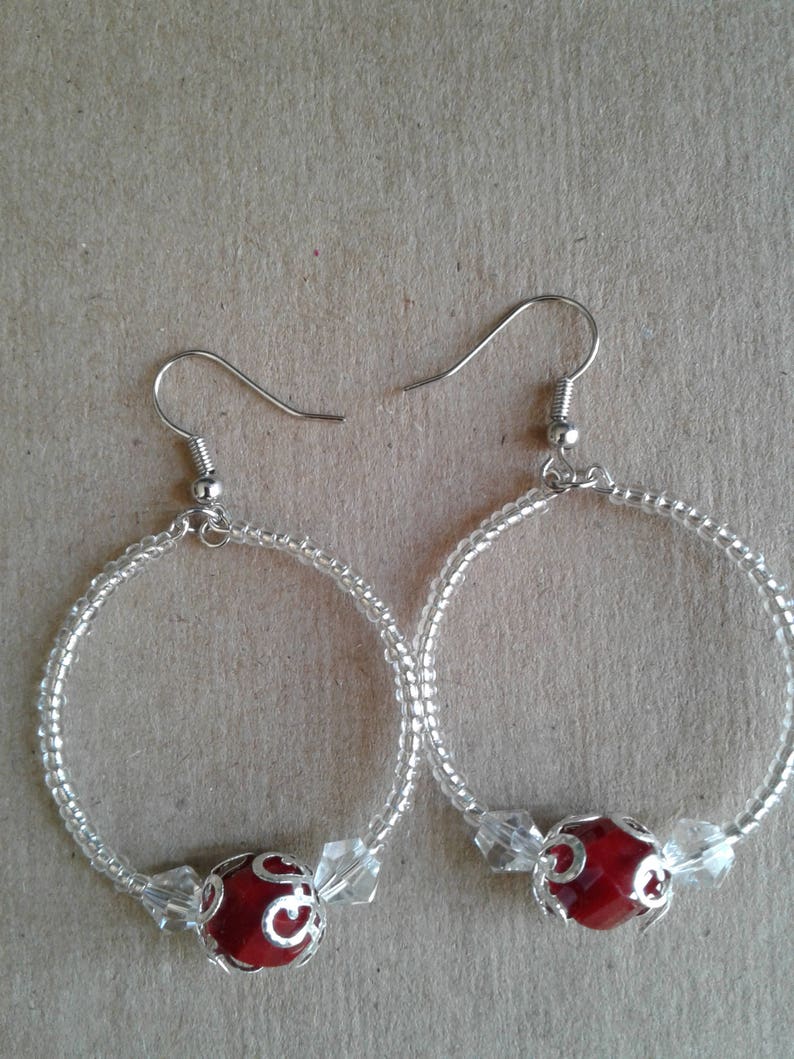 May include: A pair of silver hoop earrings with a red bead and clear crystal accents.