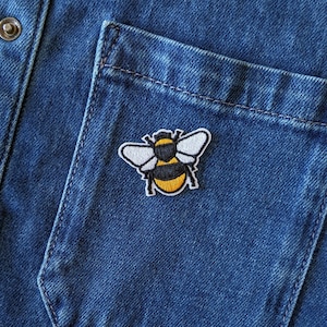 May include: A close-up of a blue denim shirt pocket featuring a yellow, black, and white embroidered bee patch. The bee patch is positioned on the pocket, with the shirt's button and seams visible.