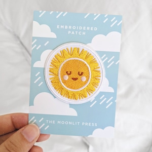 May include: A white card with blue clouds and rain printed on it. The card says "Embroidered Patch" and "The Moonlit Press". A yellow embroidered sun with a smiling face is attached to the card.