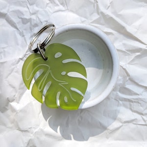 May include: A green monstera leaf keyring with a silver key ring. The keyring is sitting on a white surface.