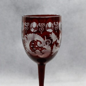 Op de afbeelding: Een rood wijnglas met een wit gegraveerd ontwerp van bloemen en krulvormen. Het glas heeft een lange steel en een ronde voet.